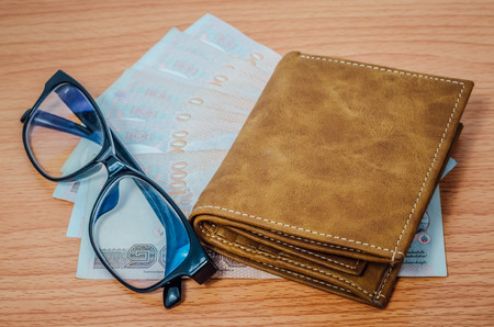 Thai money in brown leather wallet and black spectacle eyeglasses on wooden texture backgroundの写真素材