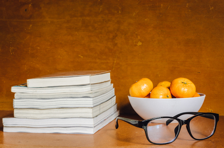 Group of oranges in white bowl, black glasses and books above wooden table and backgroundの写真素材