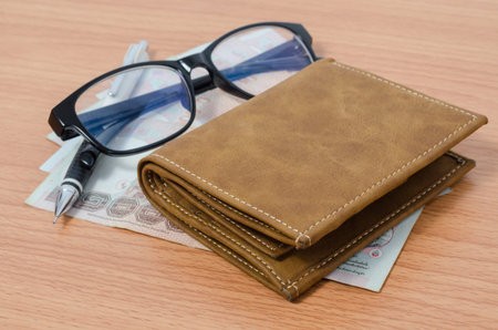 Black eyeglasses, thai money, pencil, and brown leather wallet above wooden table texture backgroundの写真素材