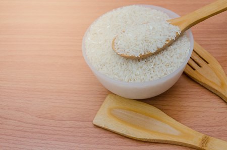 Uncooked jasmine rice grain on wooden spoon, fork, and spade with plastic bowl on wooden table background with sun lightの写真素材