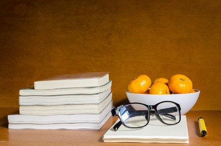 Group of oranges in white bowl, black glasses, yellow pen and books above wooden table and backgroundの写真素材