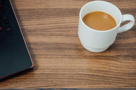 Hot coffee in white cup and black laptop above wooden table background