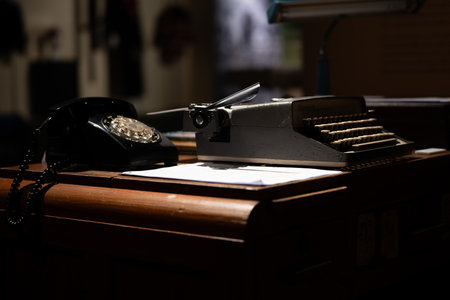 An old-fashioned black telephone and a classic typewriter rest on a wooden desk, illuminated by soft lighting, evoking a sense of nostalgia in a historic setting.の写真素材