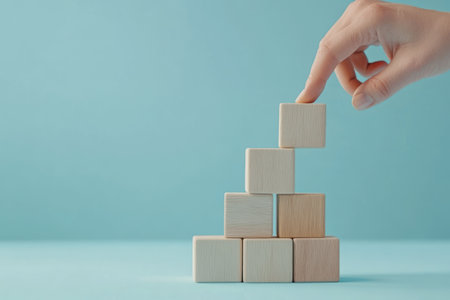 Hand stacking wooden blocks into a step formation with an arrow pointing upwards, representing the process of achieving business success, with a minimalist blue backdrop and plenty of space for text.の素材