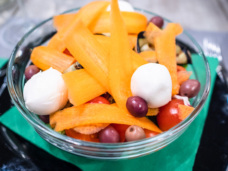 Italian cuisine - Insalata Ciliegina (salad from cherry tomatoes) with sliced carrot, mozzarella cheese and olives close up in glass bowl in local restaurant in Lombardyの写真素材