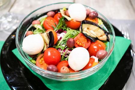 Italian cuisine - Insalata Ciliegina (salad from cherry tomatoes) with eggplant, mozzarella cheese, olives and arugula, lettuce in glass bowl in local restaurant in Lombardyの写真素材