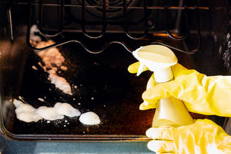 cleaning oven in home kitchen - hands in gloves spray a reagent on dirty bottom of ovenの写真素材