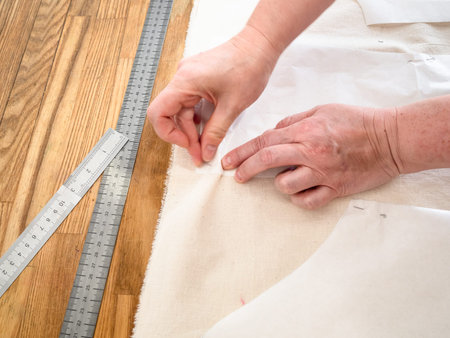 designer's hand fixes paper cutting layouts of dress to calico fabric by pins on wooden table at...