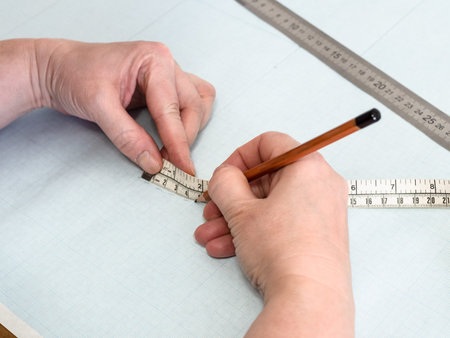 designer's hands draws pattern layout of dress with measure tape on graph paper at home