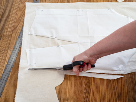 tailor's hand cuts calico fabric by scissors according with pattern layouts of dress on wooden table...