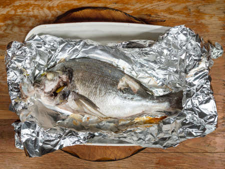 top view of Orata fish baked in foil in ceramic tray on old wooden tableの写真素材