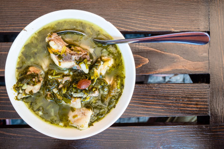top view of typical Georgian Chakapuli stew (from lamb meat, tarragon leaves, unripe plum, fresh herbs, onion, garlic, salt, wine) in white bowl on wooden table in local cafe in Borjomi town, Georgiaの写真素材