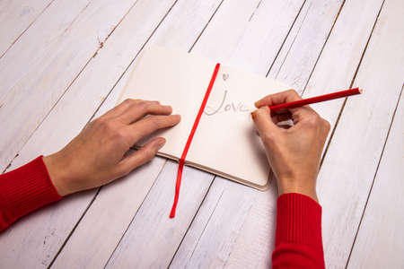 Female hands writing a lovely message on her notebook. The touch of red from the sweater and the pencil recall the Valentine color. White wooden background with empty space for text.の写真素材