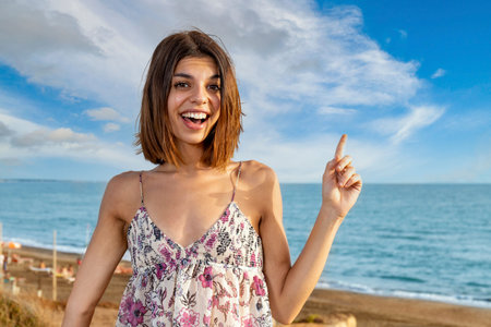 Young brunette with a surprise expression pointing her left hand up as to indicate something special. Seaside landscape in the background.の写真素材