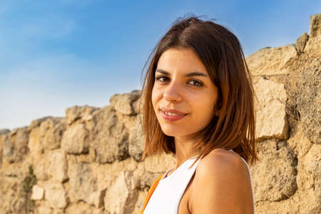 Smiling young brunette with an intense look turning her back to a rough stones wall. Bright summer colors and relaxing atmosphereの写真素材