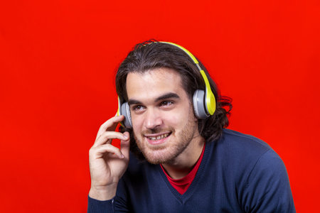Young smiling man isolated on a red background listening to music with bright yellow headphones....