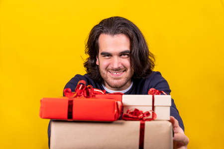 A long-haired young man distributes Christmas presents, packages with red, brown paper and red ribbon. Yellow background and copy space for your greeting message.の写真素材