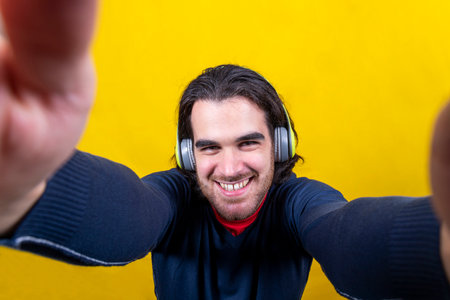 A young man with long hair and a beard is listening to music on headphones. The man is smiling while taking a selfie with his smartphoneの写真素材