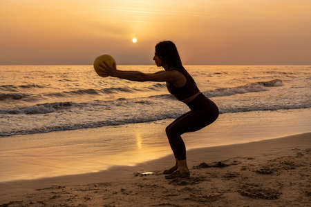 Beautiful woman doing pilates exercise on the beach at sunset. Woman doing squats with the ball. Concept of wellness and health.の写真素材