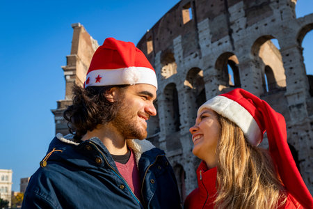Roman holidays at Christmas. A young couple poses in front of the Colosseum in red Santa Claus hats. Young people look into each other's eyes and smile. Feeling of happiness and love.の写真素材