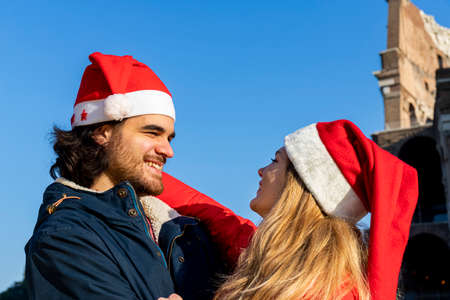 Young couple traveling to Rome. Beautiful couple in Santa Claus hat happily hugs each other in front of the Colosseum. Concept of travel, holidays, love and happiness.の写真素材