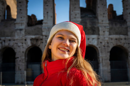 Roman holidays at Christmas. Beautiful blonde woman posing in front of the Colosseum with Santa Claus hat. The young woman looks into the camera and smiles.の写真素材