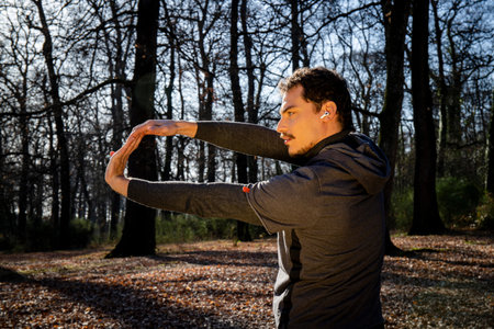 Runner is doing stretching in the woods. Young man with blue eyes and mustache is training in the...