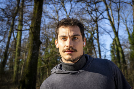 Runner is doing stretching in the woods. Young man with blue eyes and mustache is training in the...