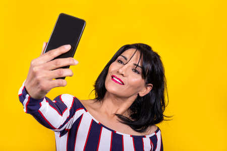 Beautiful woman is wearing a striped shirt. Young brunette woman is taking a selfie with her mobile phone on a yellow background.の写真素材