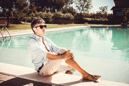 The young man is wearing sunglasses, a white shirt and beige trousers and is sitting by the pool. Handsome businessman takes a relaxing moment by the pool.の写真素材