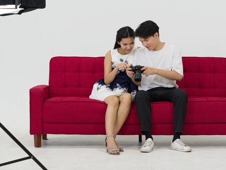 Asian photographers allow models to view pictures taken on the camera screen while sitting on a red sofa. Working atmosphere in the photo studioの写真素材
