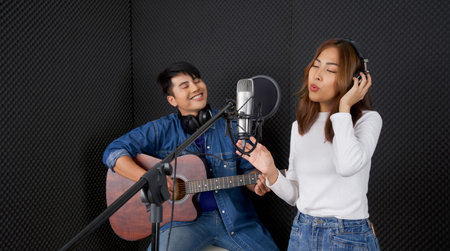 Young asian woman singing in front of black soundproofing wall. Her boyfriend played the guitar together. Musicians producing music in professional recording studio.の写真素材