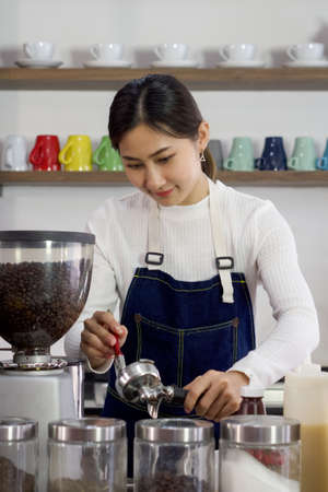 Young asian woman in apron use a brush to clean Espresso Machine Portafilters after make fresh coffee drink. Professional barista preparing coffee on counter.の写真素材