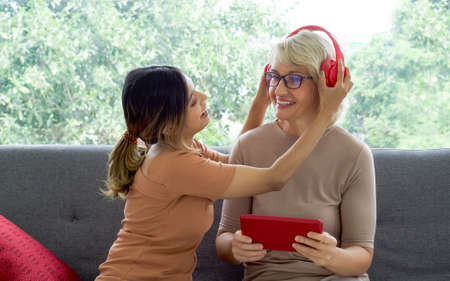 Happy mix skin family living together, asian daughter help her adopted caucasian mother listen to music with headphones through a tablet computer.の写真素材