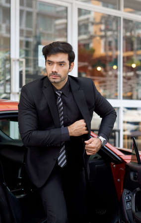 Young business man in a black suit steps down from a bright sports car while put his belonging into his suit pocket.の写真素材