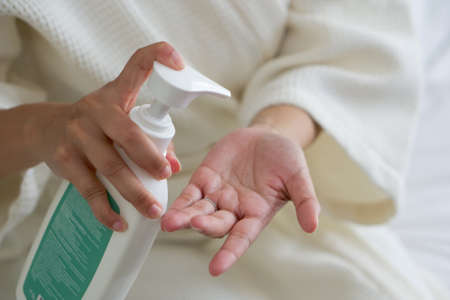 Young woman in a bathrobe presses a bottle of body lotion onto her hand while sitting in bed. Close upの写真素材