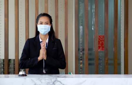The hotel receptionist raise her hand to pay respect in thai culture while wearing a surgical mask....