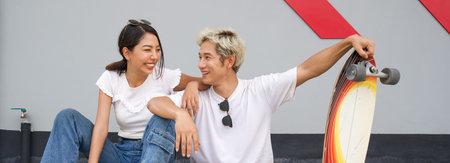 Asian couple resting after skateboarding. A young man in a white T-shirt held his hand on the surfskates board while talking to his girlfriend. The woman laughed happily with sunglasses on her head.の写真素材