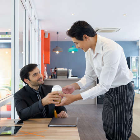 A Persian businessman in a black suit smile while receiving iced coffee from the waiter. Atmosphere during lunch break in a modern cafe.の写真素材