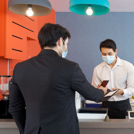 The cashier in the apron and face mask hands a receipt to a caucasian customer. A young businessman in a suit pay a service fee through Mobile applications.の写真素材
