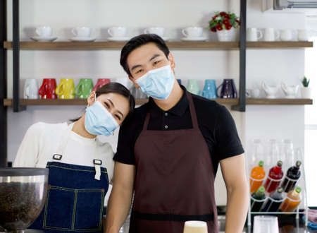 The shopkeeper and barista, wearing a face mask, poses happily before starting daily work. Morning atmosphere in a coffee shop.の写真素材