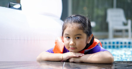 Asian child playing in the pool. Wearing orange life jacket, rests her chin on both hands by the edge of the pool.の写真素材