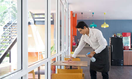 Asian waiter in an apron uses a liquid cleaner and a towel to wipe off the stains on the table. Morning atmosphere in the cafeteria.の写真素材