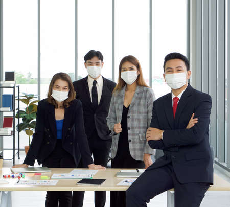 Asian businessman in a suit leaning against the table with a confident expression. The team is preparing for the meeting in the background. Everyone wears a mask. Concept of leadership.の写真素材