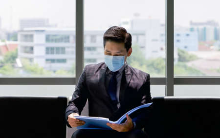 Asian business man in a suit and face mask sit on a bench near the window, examines his resume while waiting for the interview.の写真素材