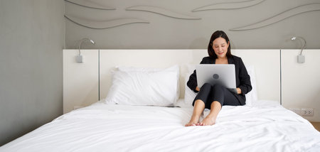 Asian woman in black suit use the laptop computer to work in the morning in bed.