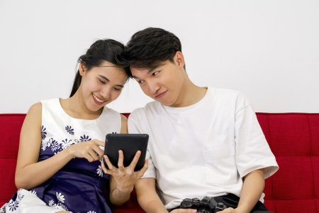 Young lovers spend time together on holidays in the photo studio. The young man allow her girlfriend to view pictures taken on tablet computer screen while relaxing on a red sofa.の写真素材