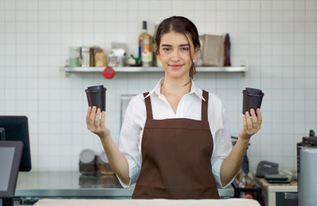 Young caucasian barista dressed in apron hold coffee drink in plastic cup with both hands in front...