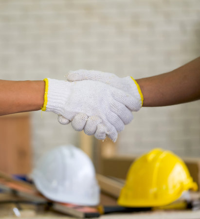 Carpenters with protective glove shaking hands after finish the job. A white and yellow construction helmet was placed on the table in the background.の写真素材