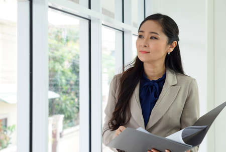 Asian female office manager with brown suit holding document folder in her hand. She looked outside while thinking of something.の写真素材